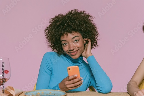 Smiling woman with curly hair holding a smartphone at a dinner table, displaying a gentle glamorous mood for social and mobile lifestyle use