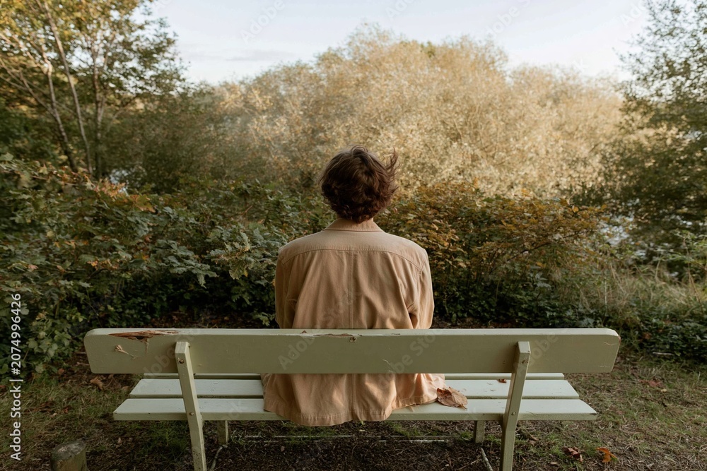 Fototapeta premium Young man seen from behind sitting on a weathered park bench gazing at autumn trees, contemplating in a peaceful nature setting for mindfulness and solitude