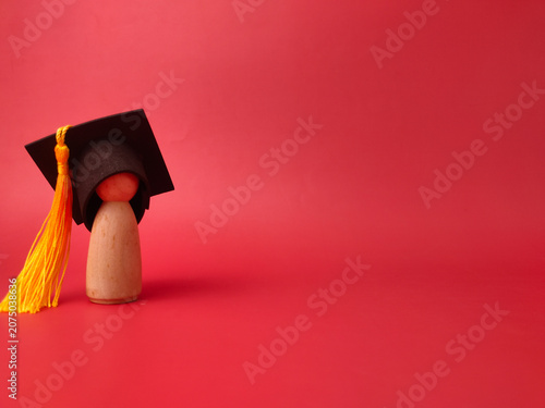 A minimal wooden peg figure wears a black graduation cap with a long yellow tassel against red