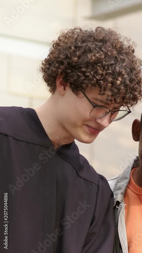 Vertical video: Turning grad in robe specs, friend in jean jacket showing phone at stone wall