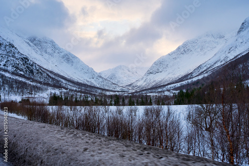 Majestic Winter Road Trip in Lofoten: Majestic Snow-Covered Mountains and Distant Cabin by Frozen Fjord Under Moody Cloudy Sky, Scenic Nordic Landscape in Norway.