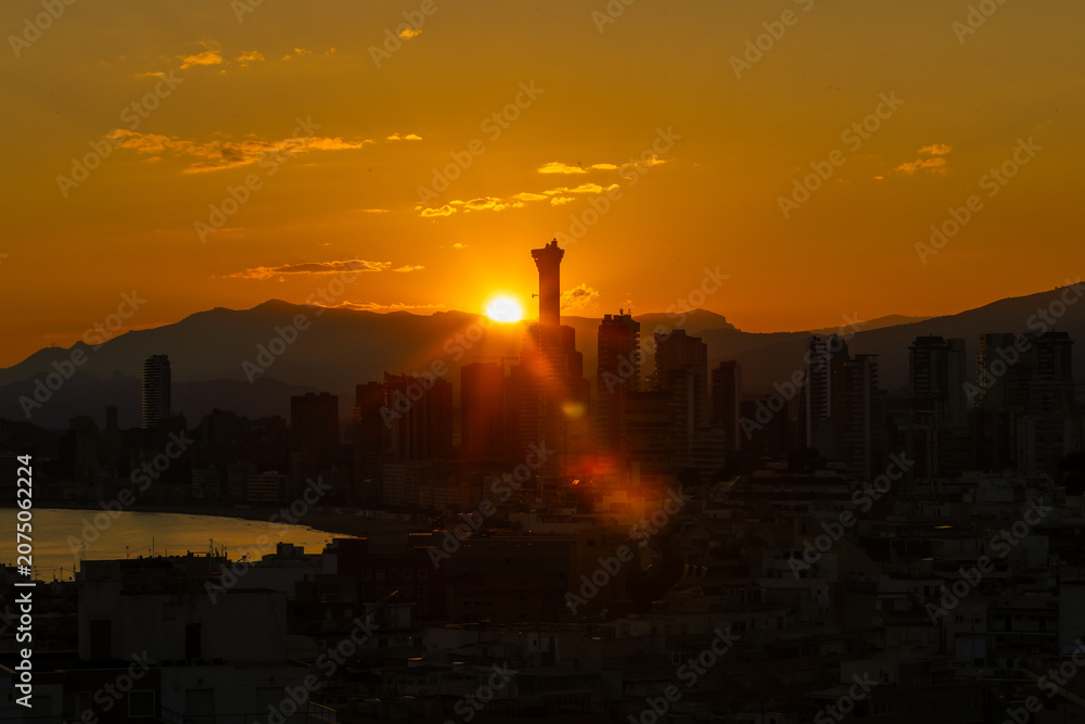 Fototapeta premium Dramatic sunset over the Benidorm skyline in Spain with the Intempo Building silhouetted against a glowing orange sky and distant mountains creating a striking cityscape above the Mediterranean coast