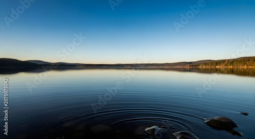 Calm lake with ripples and rocks, reflecting the sky and distant hills, serene nature landscape