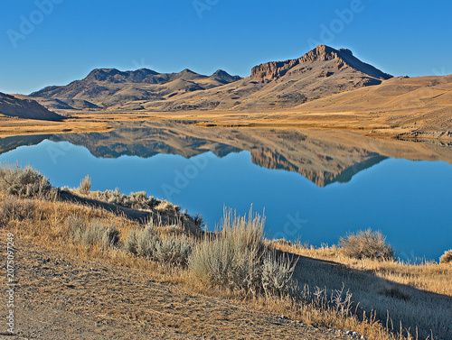 Mountains reflecting in a serene blue lake, framed by dry golden hills and foreground brush under a clear sky
