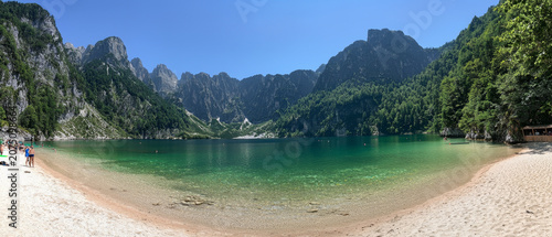 Panoramic vista: clear emerald lake with sandy beach and people, surrounded by majestic, forested mountains under a blue sky