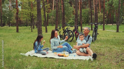 Happy family resting on blanket in forest park eating fruits after bike ride with bicycles nearby enjoying outdoor time together in nature