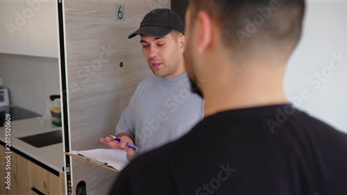 Delivery worker wearing gray t-shirt and black cap holds clipboard while speaking with customer at modern apartment doorway number six, taking notes during friendly doorstep conversation.