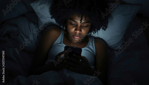 Black young woman with a worried expression uses her mobile phone at night in bed, reading a text message with bad news, experiencing stress and sadness at home