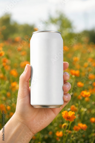 Hand holding a blank white aluminum beverage can mockup in front of a beautiful, blurred field of bright orange flowers under daylight.