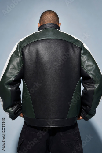 Rear view of a man wearing a stylish black and dark green leather varsity jacket with white stripe accents, standing against a blue studio backdrop