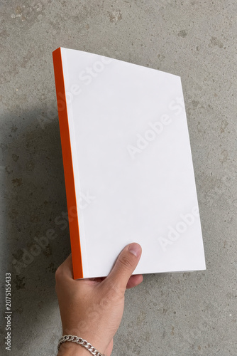 Hand Holding Blank White Book Mockup with Orange Spine Against Concrete Background