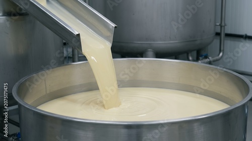 Creamy pale liquid is poured from a metal chute into a large stainless steel industrial vat creating ripples on its surface pouring food production dairy manufacturing tank