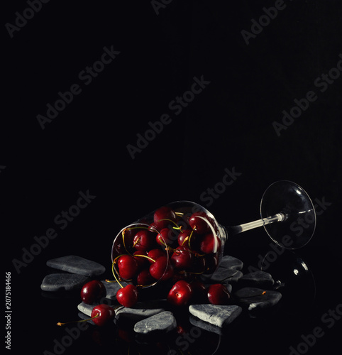 Still life. An overturned glass, scattered sweet cherries, and stones. Set against a black background with a reflection.