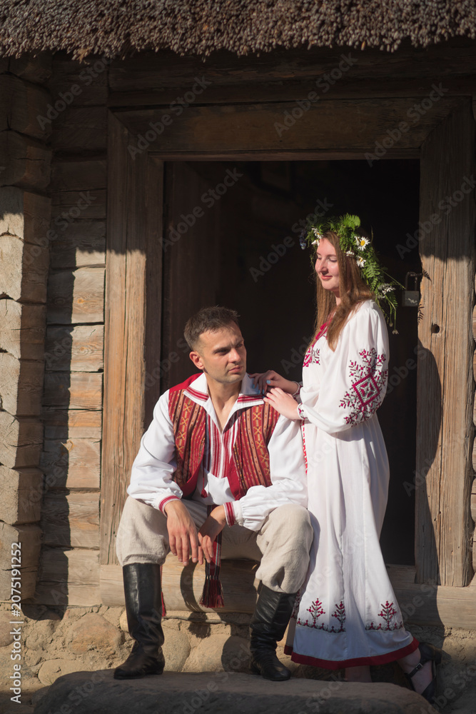 Fototapeta premium Romantic slavic couple in love wearing traditional embroidered clothes by wooden house with copy space