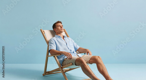 Man sitting in wooden deck chair against blue background. Young guy relaxing during summer vacation. Leisure time and rest concept. Minimalist studio studio shot of traveler guy.