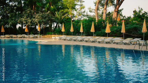 Outdoor swimming pool with blue water, empty sun loungers and yellow striped umbrellas. Luxury resort hotel relaxation zone, summer vacation concept.