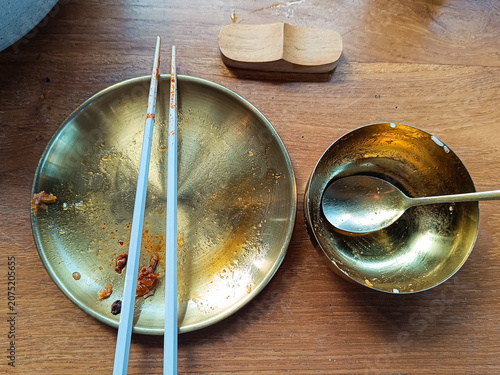 empty golden plate with chopsticks and a small bowl on a wooden table after a meal. The leftover food pieces and dining utensils create a casual restaurant atmosphere with warm lighting and a cozy