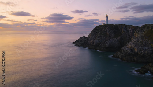 Peaceful lighthouse standing on a rocky coastline overlooking calm ocean waters during a colorful sunset, offering a serene coastal landscape.