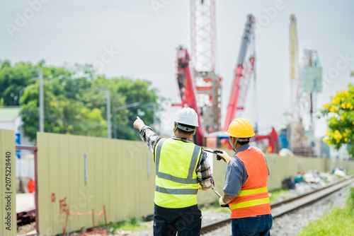 Construction professionals inspecting site progress. Two engineers in safety gear point towards urban infrastructure development, emphasizing project management and teamwork.