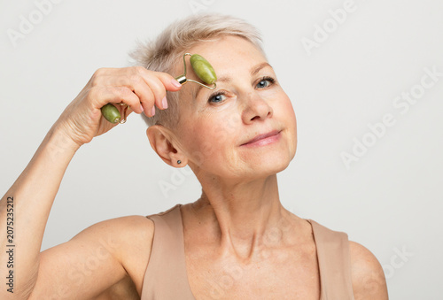Gorgeous woman of 60s massaging face skin using natural facial stone trendy jade green quartz roller