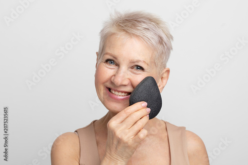 Charming elderly woman with short hair use sponge remove make up on light grey background.