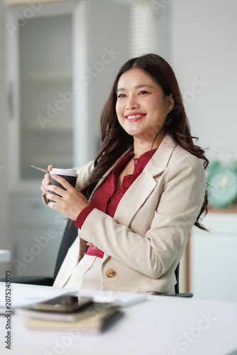 Asian businesswoman smiling and holding a coffee cup, taking a break from work in an office, symbolizing successful career and professional female leadership in business