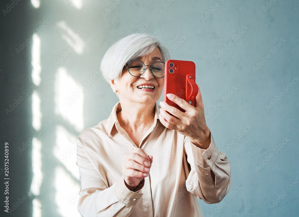 Fototapeta premium An elderly woman with short hair smiles softly while holding up her red phone, enjoying a lighthearted selfie moment.