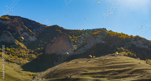 Paragliders soar gracefully against rugged mountain backdrop, with vibrant autumn foliage below and clear blue sky above. Fly Chegem paragliding site, El-Tyubu, Russia - October 7, 2024