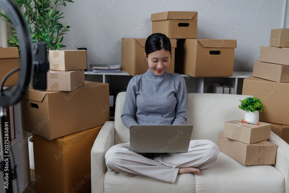 Fototapeta premium Young asian woman sitting on a sofa in a home office, using a laptop surrounded by numerous cardboard boxes, efficiently managing her growing online e-commerce business
