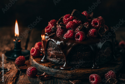 Dark moody still life of a decadent chocolate cake with dripping glaze topped with fresh raspberries and chocolate curls illuminated by a single burning black candle on a rustic wooden board
