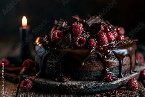 Dark moody still life of a decadent chocolate cake with dripping glaze topped with fresh raspberries and chocolate curls illuminated by a single burning black candle on a rustic wooden board