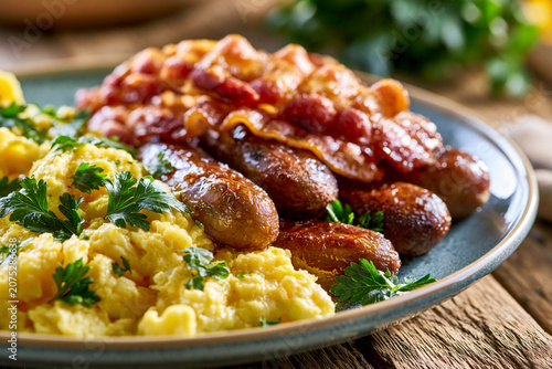 Delicious traditional english breakfast plate with fluffy scrambled eggs garnished with fresh parsley, crispy smoked bacon strips, and juicy grilled sausages on a rustic wooden table