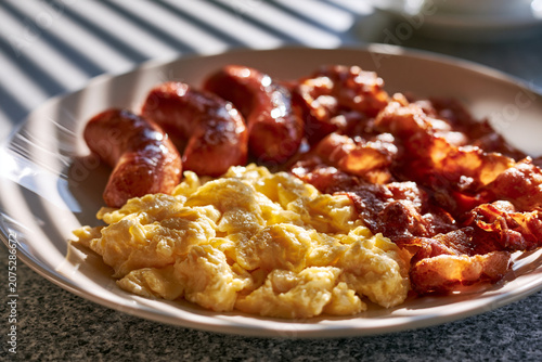 Delicious traditional english breakfast plate with fluffy scrambled eggs garnished with fresh parsley, crispy smoked bacon strips, and juicy grilled sausages on a rustic wooden table