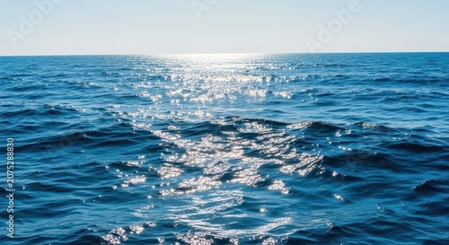 Deep blue ocean surface stretching toward the horizon under a bright clear sky. Sunlight sparkles on the choppy water and small rolling waves, fluid, background, wide
