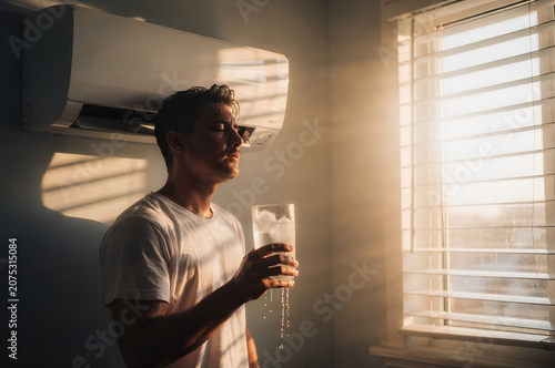 Sweaty young man drinking a glass of cold ice water while cooling off under an air conditioner during a summer heatwave
