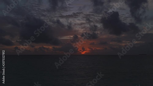 Aerial wide shot at dusk shows Maldives open water, a tiny atoll at far right, heavy cumulus and rippled clouds, orange sunset band, drifting clouds, gentle sea ripples.