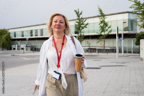 Senior female doctor wearing a white coat and an id badge, holding a reusable cup and a smartphone, walking confidently on a sidewalk outside a modern hospital building