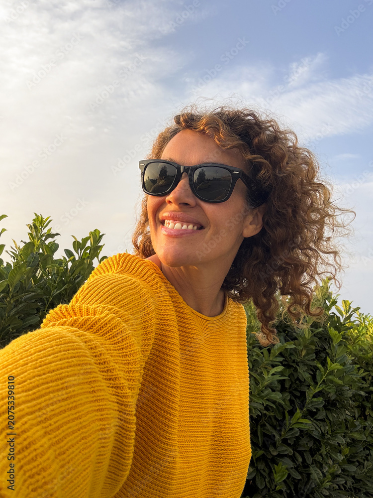Fototapeta premium Smiling woman taking a selfie outdoors wearing sunglasses and a yellow sweater with curly hair against green foliage and a blue sky