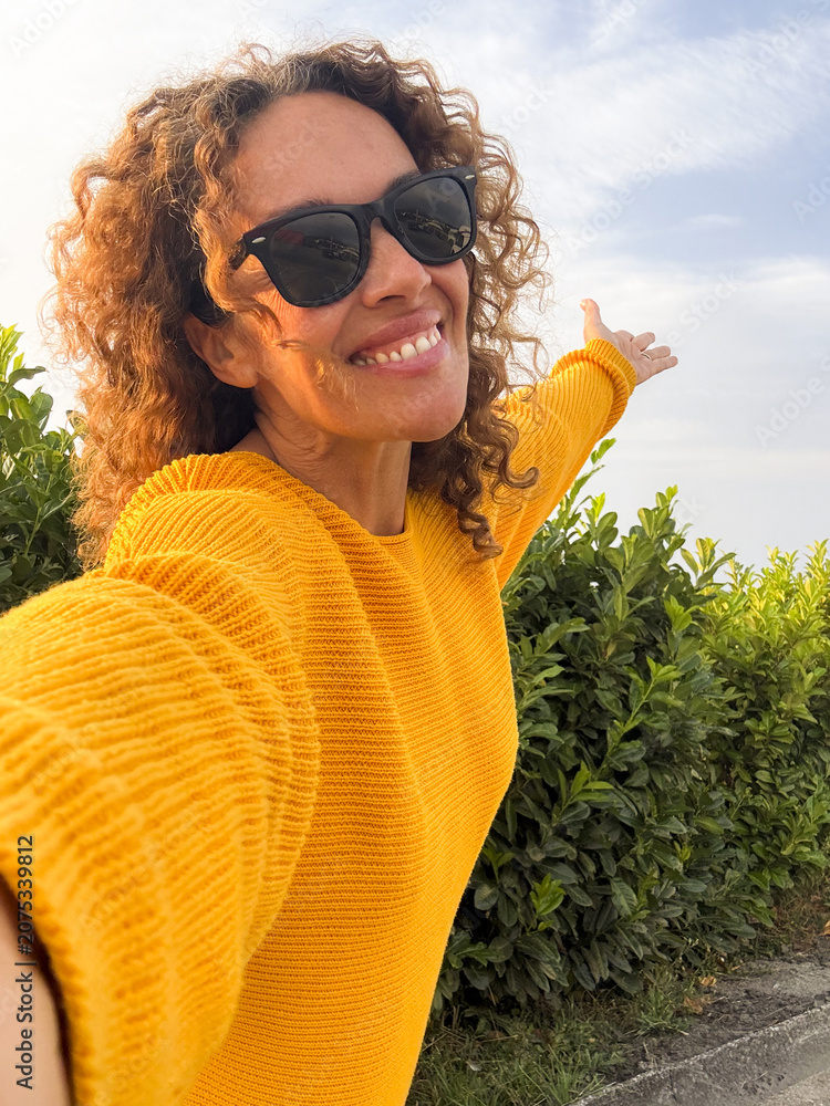 Fototapeta premium A smiling woman in a yellow sweater takes an outdoor selfie with sunglasses, arms outstretched, enjoying sunshine and nature near green bushes