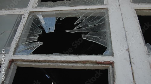 Close-up of a shattered window in an old abandoned building, revealing jagged glass fragments and weathered wooden frames. The damaged pane creates a dramatic urban decay scene, symbolizing neglect