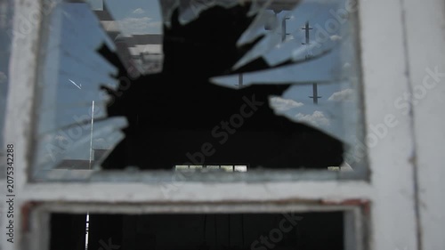 Close-up of a shattered window in an old abandoned building, revealing jagged glass fragments and weathered wooden frames. The damaged pane creates a dramatic urban decay scene, symbolizing neglect