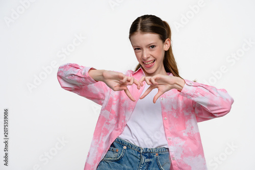 Teenage girl forming a heart shape with her hands and smiling. Studio portrait on light background expressing love, care and positive emotions.