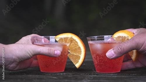 Slow motion two hands placing refreshing red cocktails with orange slices and ice cubes on a rustic wooden table. People toasting for a celebration or party