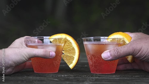 Two hands picking up plastic cups with orange-garnished cocktails from a wooden table. Friends making a celebratory toast outdoors