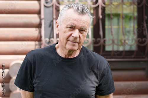 Senior man in a black t-shirt looking at camera outdoors near a wooden house, authentic lifestyle portrait with copy space.