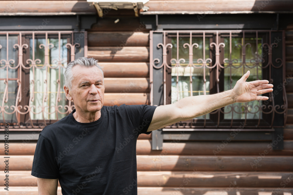 Fototapeta premium Senior man gesturing with one arm outdoors near a wooden house, concept of explaining, welcoming, guiding or presenting a place.