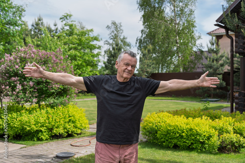 Full body portrait of a senior man standing with open arms in a green yard, symbolizing welcome, openness and life satisfaction.