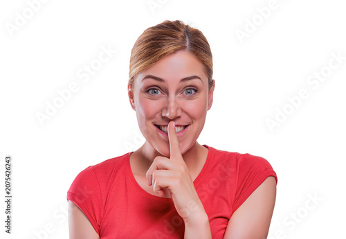 A smiling young woman in a red t-shirt holds her index finger to her lips in a shushing gesture against a white background.