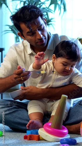 Indian father feeding food to crying baby boy showing tantrums while sitting on carpet in bright modern home, lovingly handling difficult mealtime with patience, care, and emotional parenting indoors