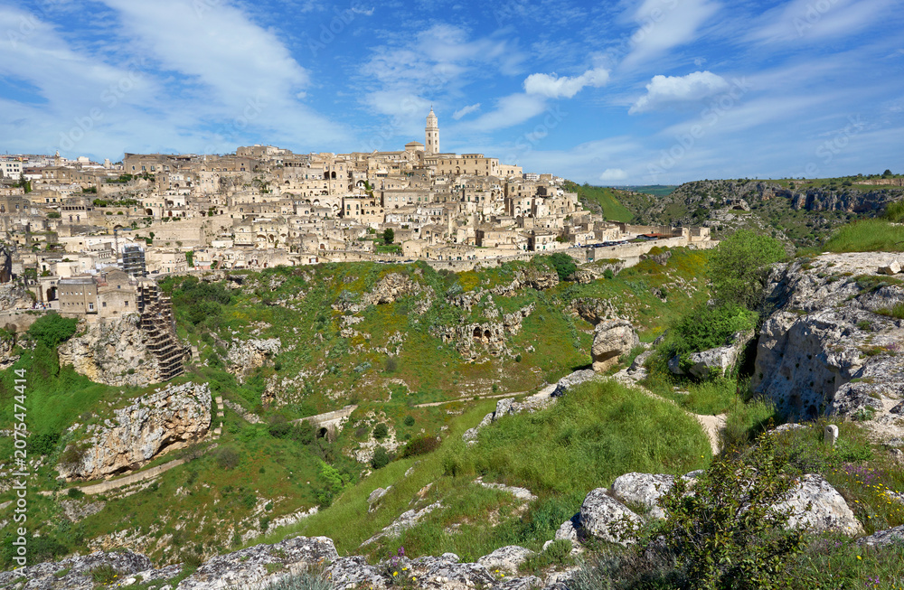 Fototapeta premium Panoramic view of the medieval cave city with historic stone architecture, dramatic cliffs and lush valley beneath a clear blue sky. Matera, Basilicata, Italy